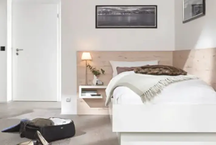 Weisses Bett mit hellen Holzpaneelen an der Wand, links schwebender Nachttisch mit kleiner Lampe und Vase, davor offene Reisetasche auf dem Boden.